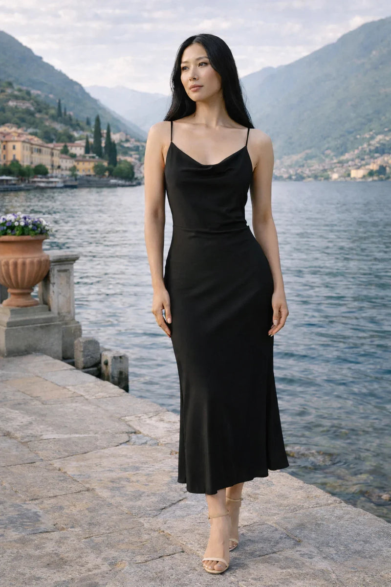Mulher usando vestido preto midi de alças finas, em cenário à beira de lago e montanhas.
