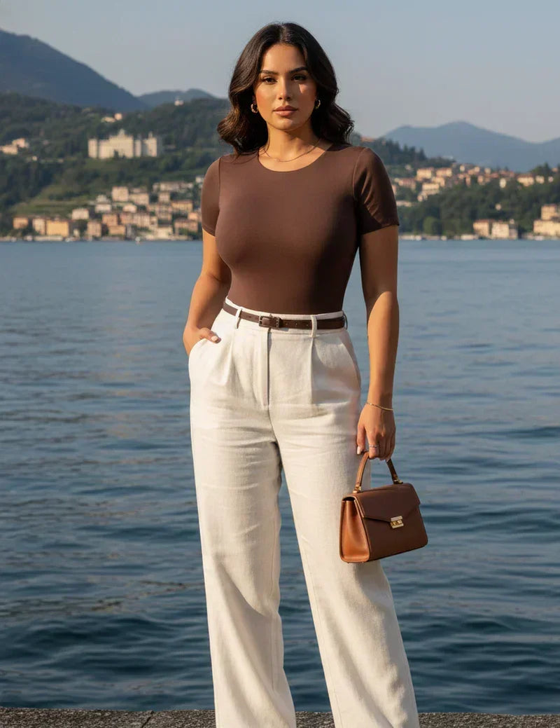 Mulher elegante com blusa marrom e calça branca, bolsa de couro, lago e montanhas ao fundo.