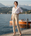 Mulher elegante com camisa branca e calça bege à beira de lago, barco e montanhas ao fundo.