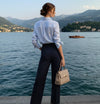 Mulher elegante com camisa listrada e calça social, segurando bolsa bege à beira de um lago.
