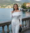 Mulher elegante com roupa branca de ombro a ombro, segurando bolsa preta, à beira de um lago.