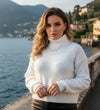 Mulher usando suéter branco de gola alta à beira de um lago, com casas e montanhas ao fundo.