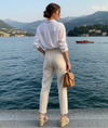 Mulher elegante de costas à beira de um lago, vestindo camisa branca, calça clara, sandálias de plataforma e bolsa marrom, com paisagem de montanhas ao fundo.