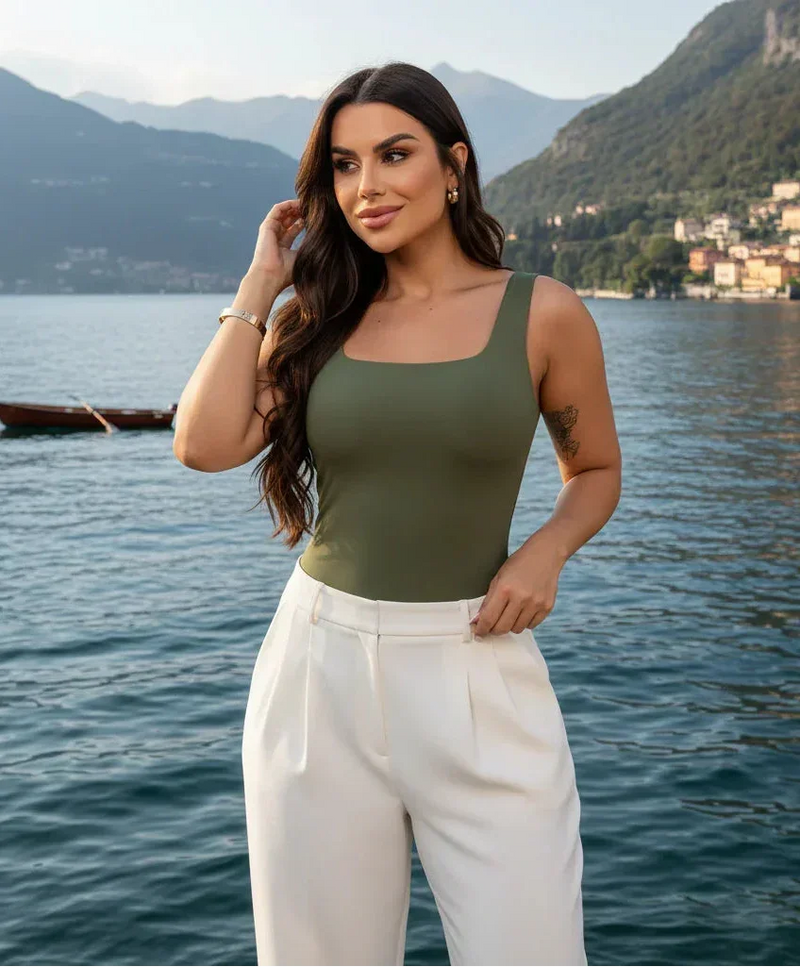 Mulher com body verde e calça branca, em frente a um lago, com montanhas ao fundo.