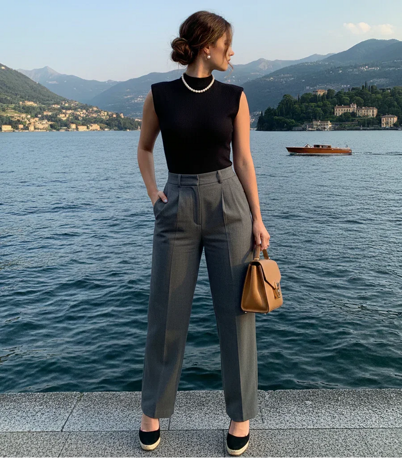Mulher elegante à beira do lago usando blusa preta sem mangas, calça social cinza e bolsa marrom.