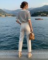 Mulher de costas à beira de um lago, vestindo calça branca, blusa listrada e bolsa marrom elegante.