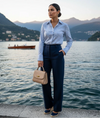 Mulher elegante com camisa listrada azul, calça social azul-marinho e bolsa bege à beira de um lago