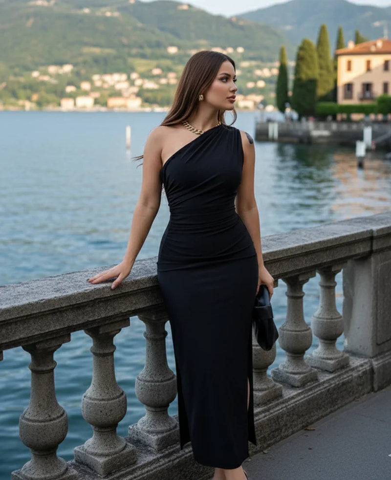 Mulher com vestido preto de um ombro só posando à beira de um lago com paisagem ao fundo