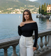 Mulher de cabelo liso com blusa preta ombro a ombro e calça branca, posando à beira de um lago com montanhas ao fundo.