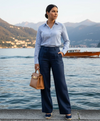Mulher elegante com camisa listrada azul e calça social azul-marinho à beira de lago, segurando bolsa marrom.